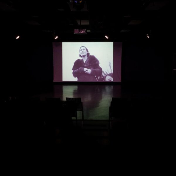 Dark Room with a film image projected in the center of a woman in a fur coat holding her hands together. In the corner a woman looks back at camera.