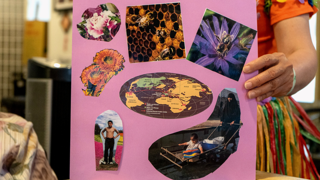 A person with orange shirt is holding a collage art on a bright pink paper. The paper shape is square and there are seven cut out images on it. From left to right; a picture of pink flower, a close-up of a beehive, a close up of a purple flower with a bee on top of it, two sunflowers, a world map, a cut-out picture of a shirtless man on another picture of a colorful flower field and a picture of a woman in an all-black outfit pushing a cart with a baby in it.