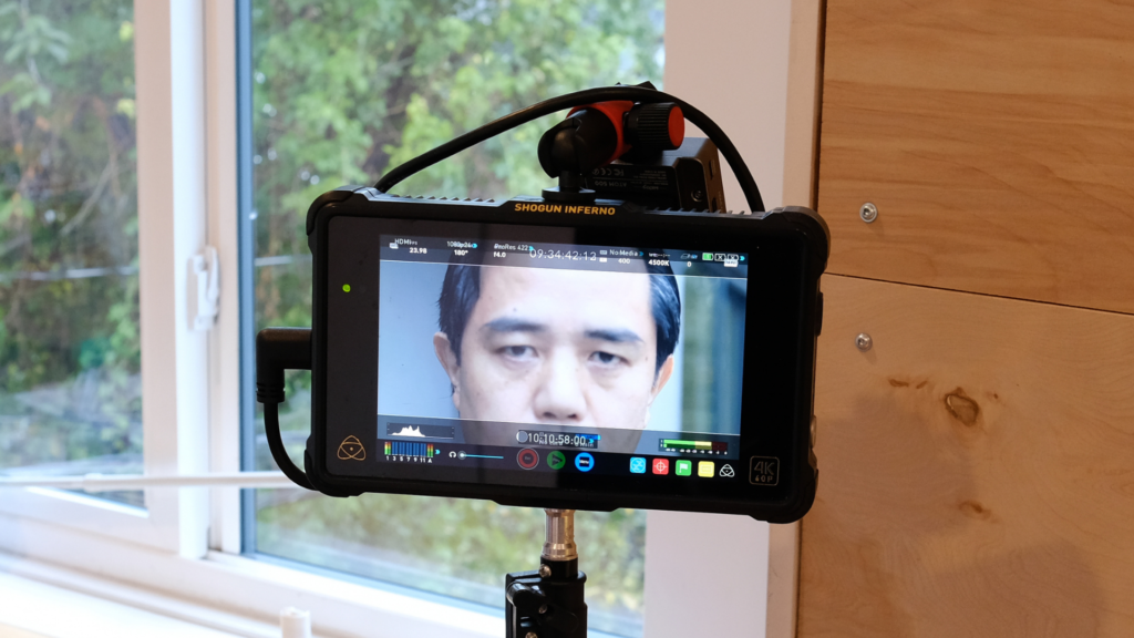 A close-up of a person's face on a screen. The person has short, dark hair and deep brown eyes that are staring directly at the camera. The screen has a black frame and it sits on a tripod. There's a window with white frame on a wood panel behind the screen that looks out to some trees.
