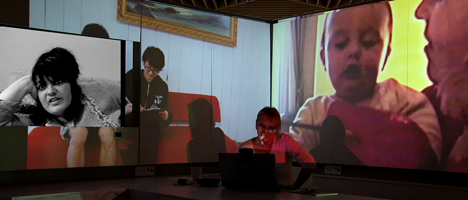 Margaret Dragu sitting with a laptop open on the table and in the background there are images of women projected onto the wall; one is talking to the camera, one is sitting on the sofa and writing and one is a close-up of a woman holding a baby.
