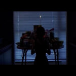 Film still from Sisters. In a darkened room, a skeletal figure stands in front of a table piled high with packaged foods. A single candle rises above the altar.