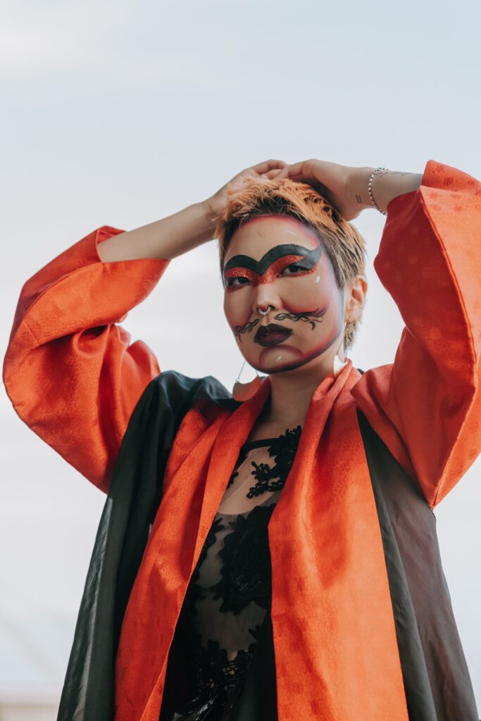 Performance photo of Skim (in drag) wearing an oragne and black cape, with heavy black and orange makeup on their face, over eyebrows , defining cheeks and chin.