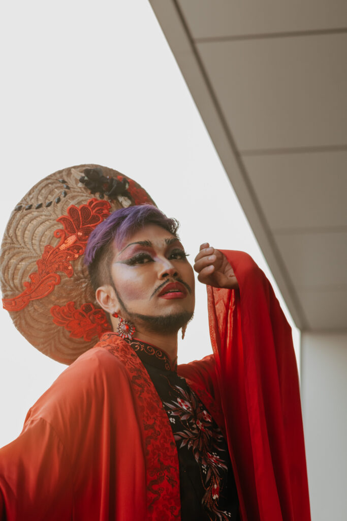 Performance photo of Shay Dior in drag costume. Shay wears a big straw hat with red decoration d a red robe.