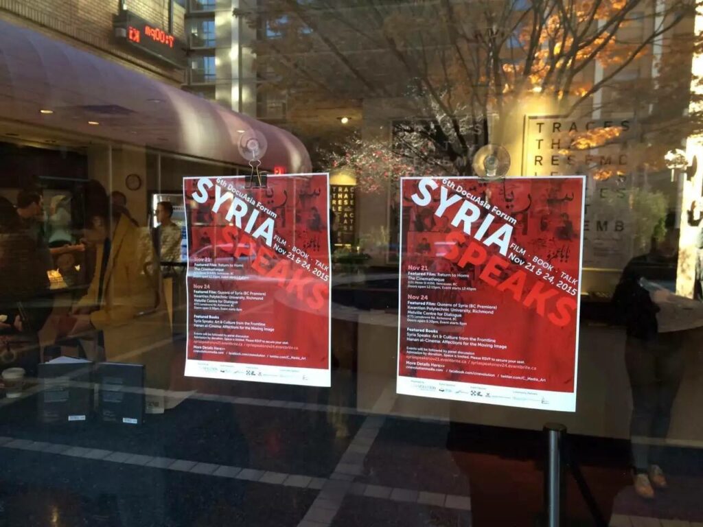 Window front with 2 posters showing the info for DocuAsisa Forum Syria. Refelections of tree from outside and people inside.
