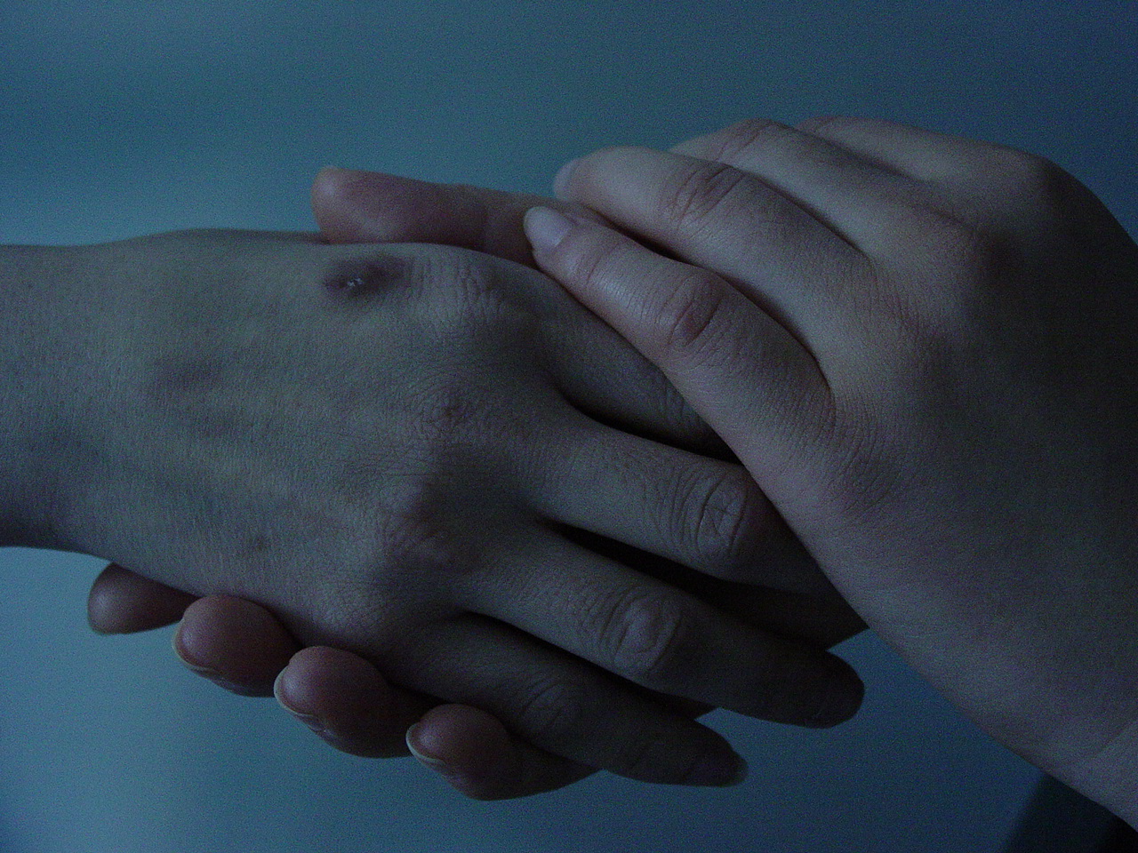 Still from Sisters. A dimly lit close-up of the hands of two people. The bruised hand of one person is held gently, supported from the bottom and the top, by both hands of the other.