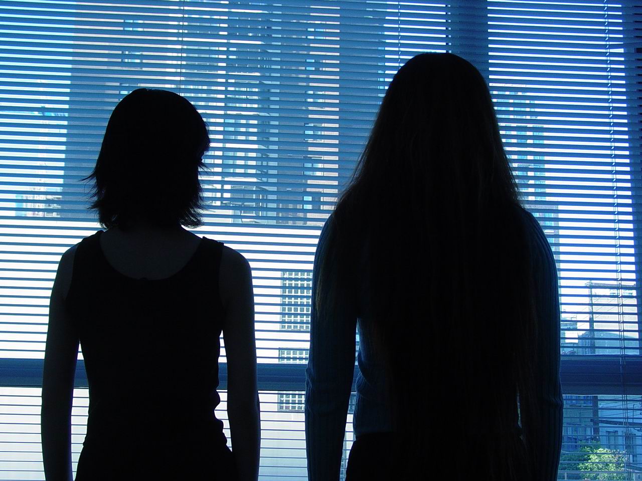 Still from Sisters. The figures of two people are shown in silhouette, looking out through Venetian blinds at the cityscape.
