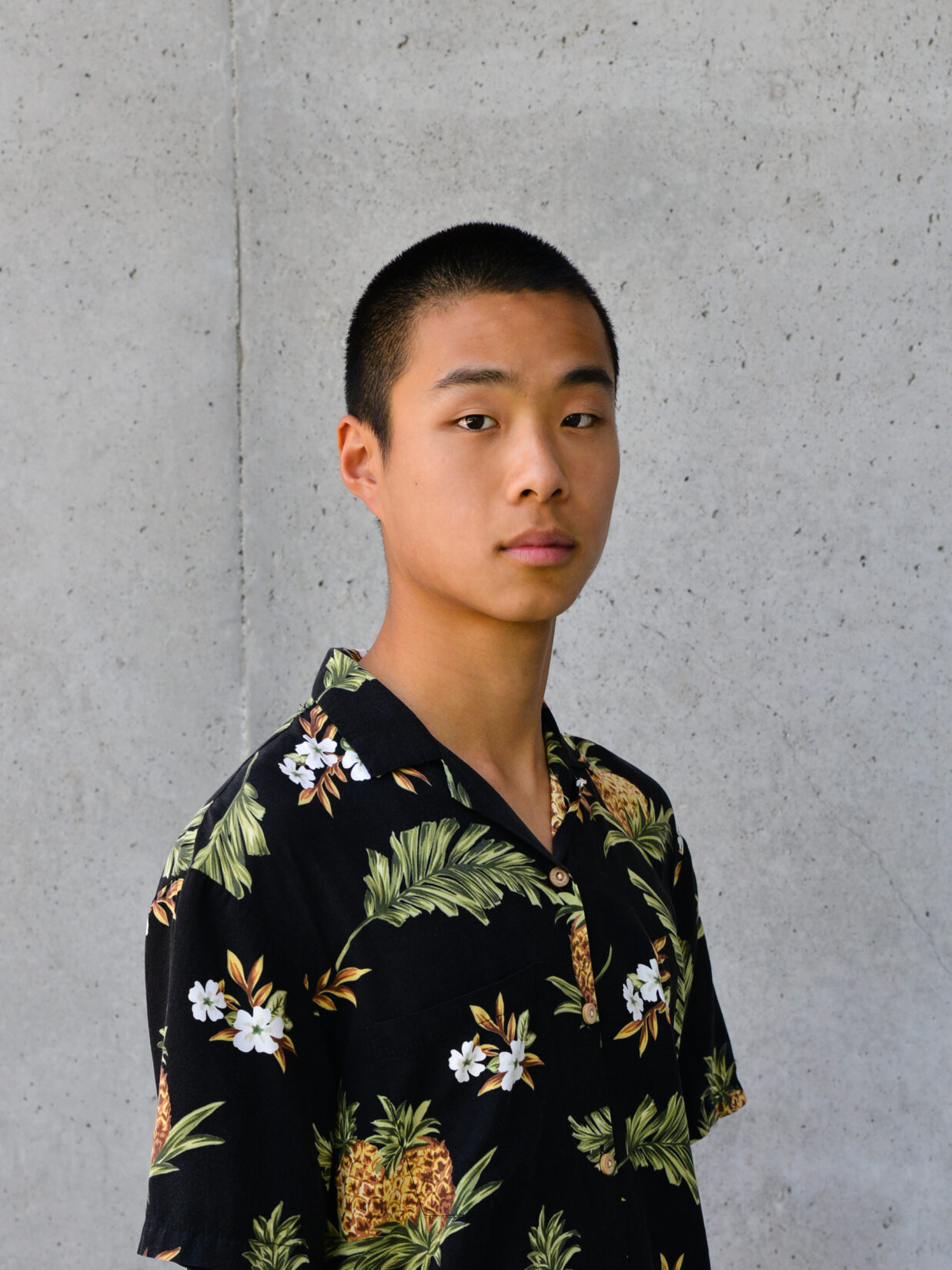 Headshot of Alger Liang. He's wearing a printed shirt which stands out starkly against the concrete wall behind him. Expressionless, there is a certain vulnerability in the length of his neck and his close cropped hair.