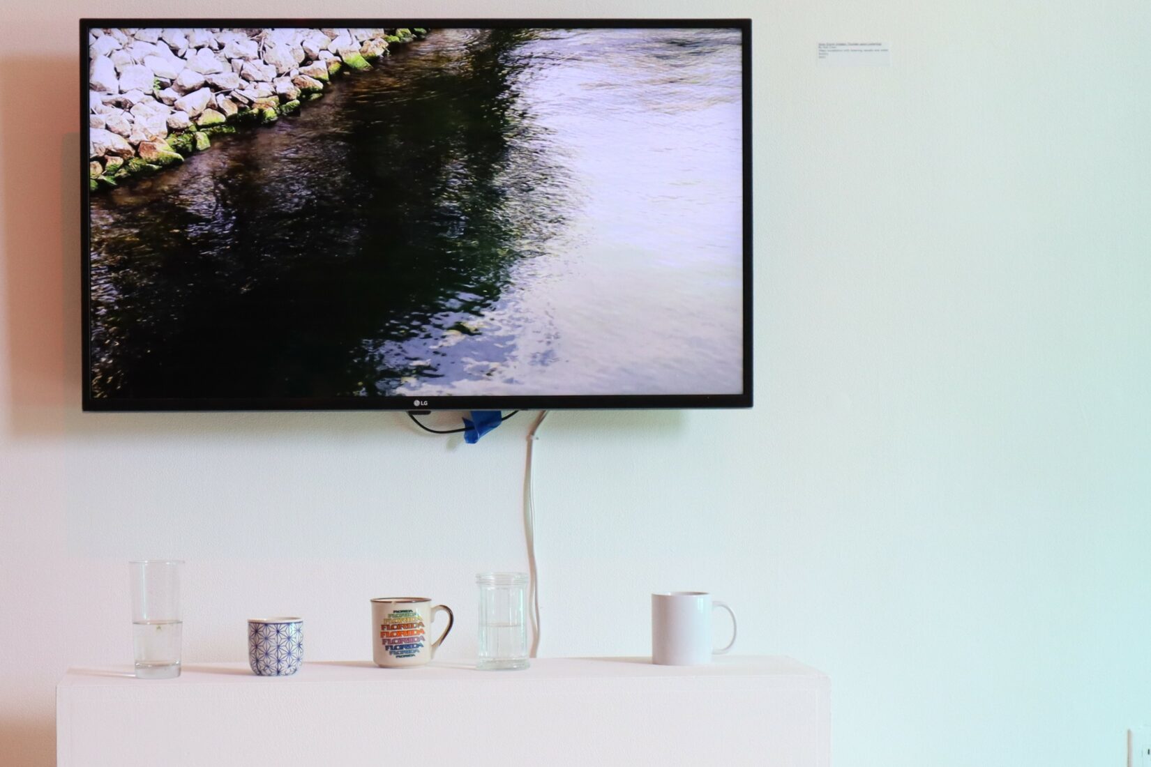 An installation photo with a large TV monitor hanging on a mint green wall, showing an image of a river with rocky shores. In front of the TV, three ceramic mugs and two glasses of different heights and colours sit on a white horizontal bench.