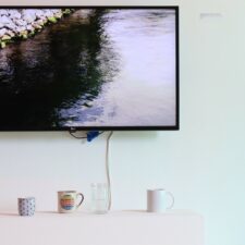 An installation photo with a large TV monitor hanging on a mint green wall, showing an image of a river with rocky shores. In front of the TV, three ceramic mugs and two glasses of different heights and colours sit on a white horizontal bench.